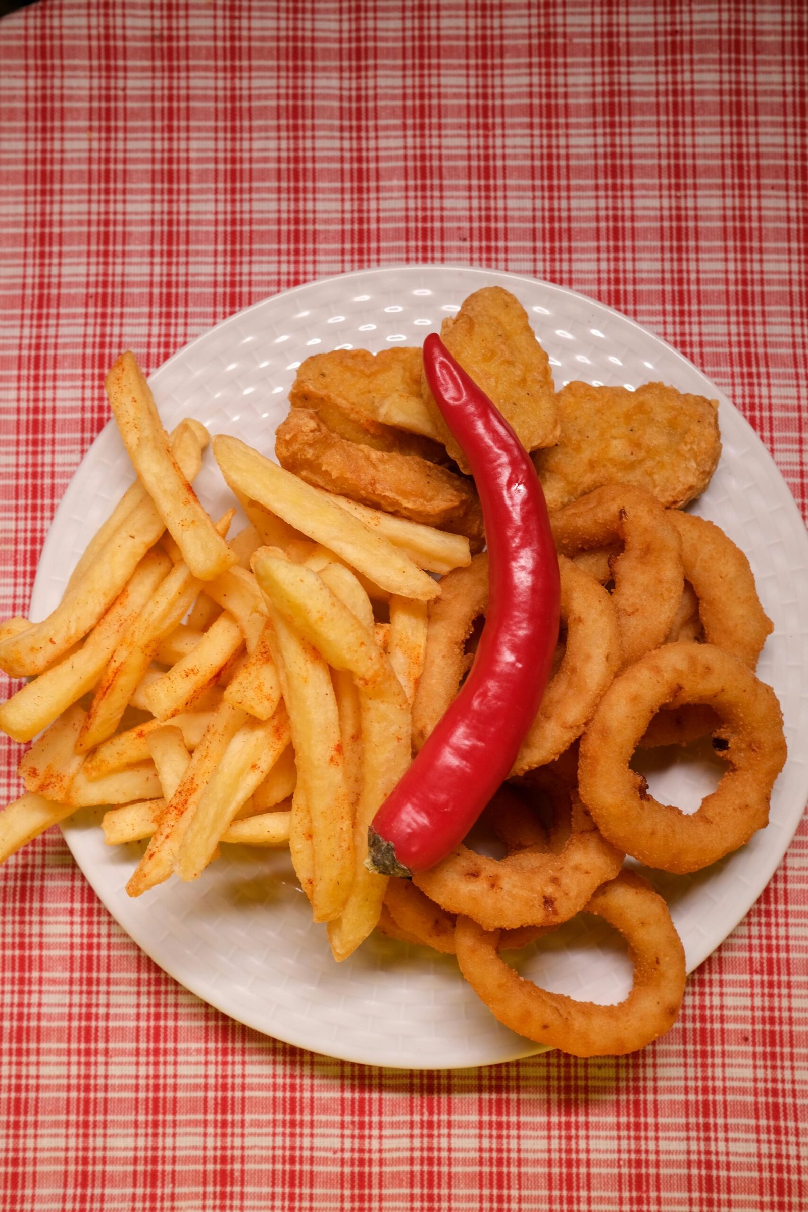 Delectable assortment of crispy snacks including fries, nuggets, and onion rings with a chili pepper on a checkered tablecloth.